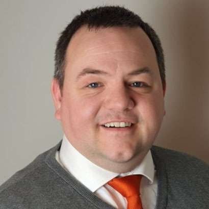 Professional headshot of a man with dark hair wearing a gray blazer, white shirt, and red tie against a neutral background